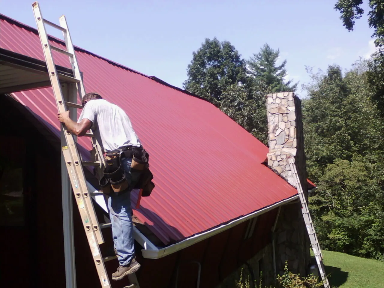 Roofer on a ladder beside a red metal roof and stone chimney — Soffit & Fascia Repair by Roofing Brothers in Russiaville